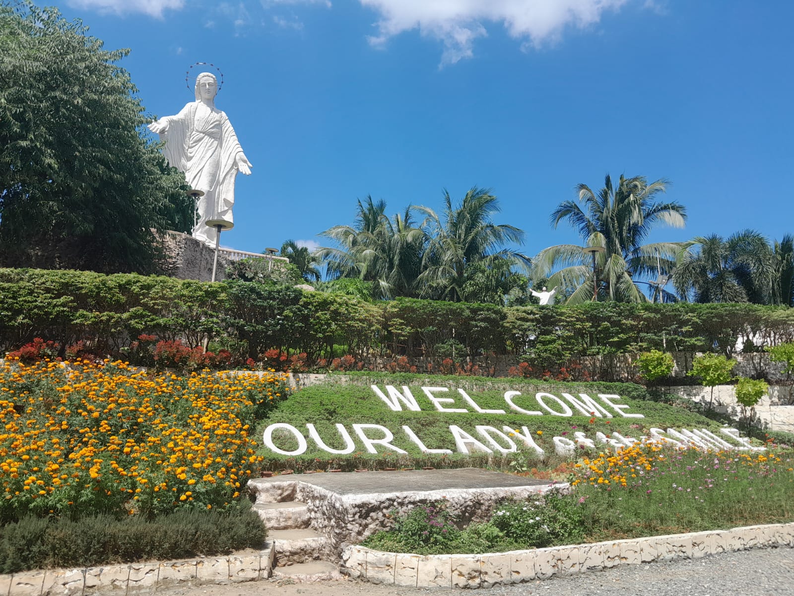 The Our Lady of the Smile, i.e., the Virgin Mary. She sits at the back of the image, towering the landscape. Below her is a signage labelled "Welcome Our Lady of the Smile" surrounded by orange and red flowers..