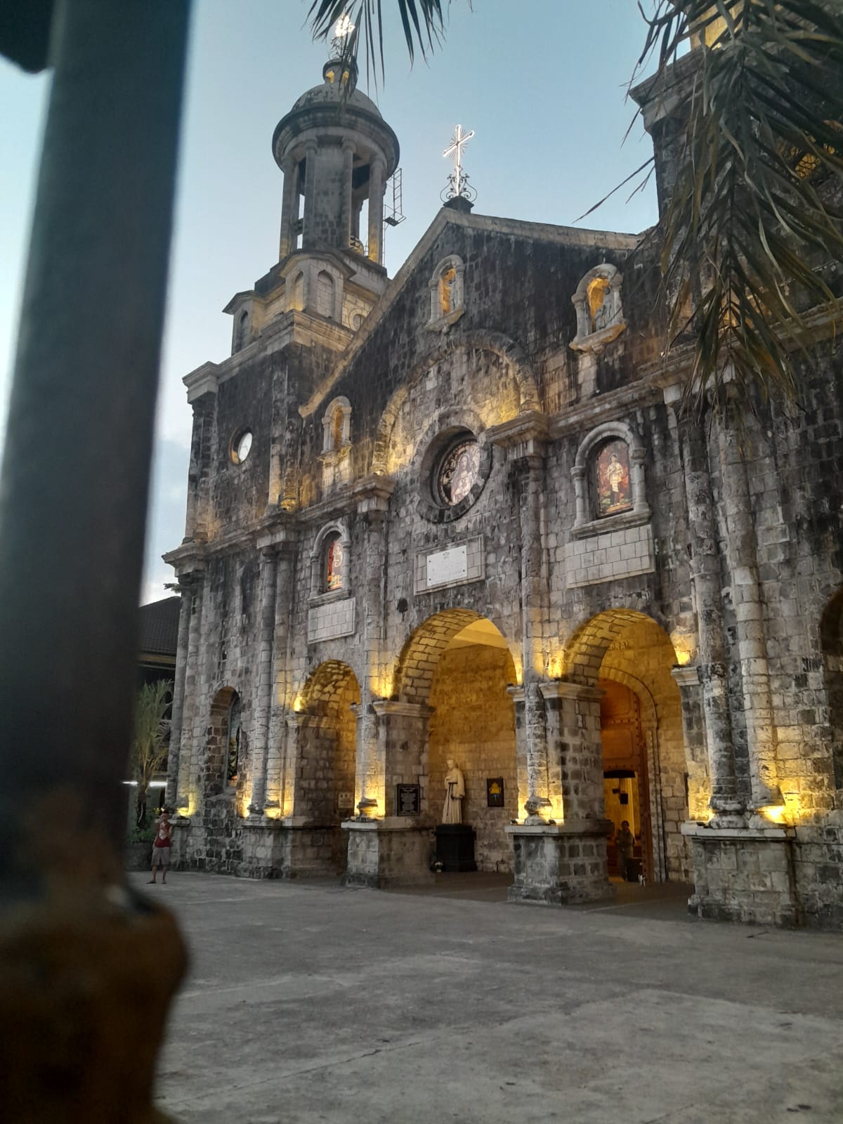 The front of San Sebastian Cathedral. Grey colored sandstone with lights shining upon its walls in the early evening. The cross at the very top also lit up.